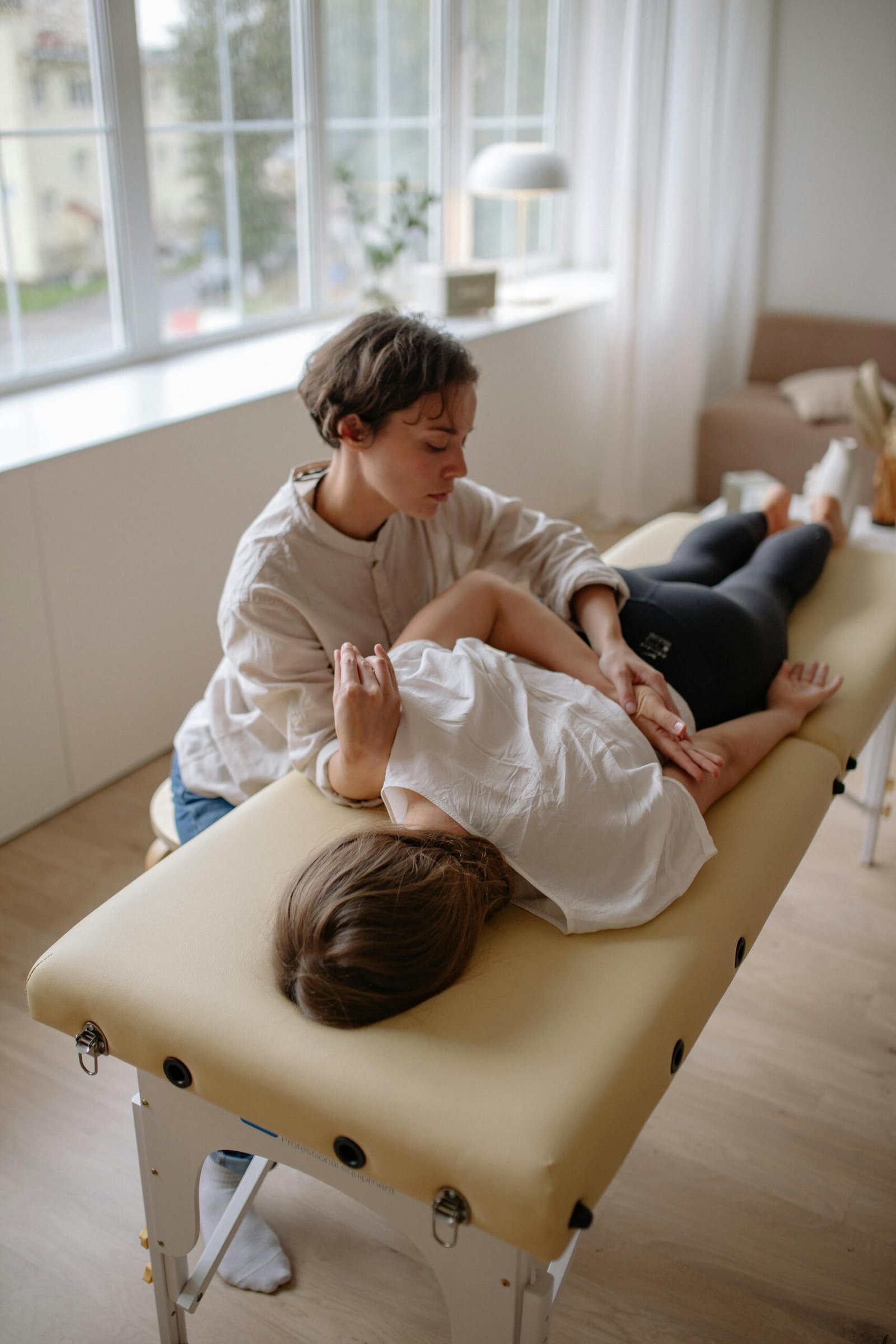 A woman lying on a massage table receiving a chiropractic adjustment in a bright room.